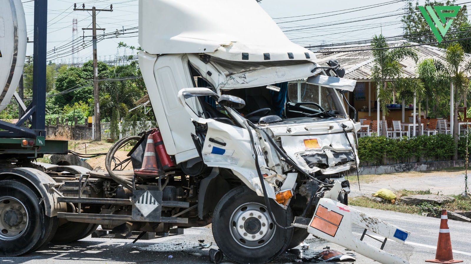 A damaged white semi-truck with a crushed cab is shown on the side of a road near a rural area with a small building in the background.