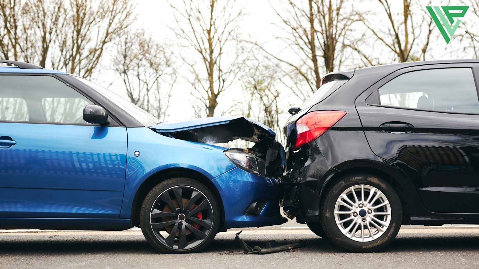 A close-up image of a rear-end collision between a blue car and a black car on a road. The blue car's front end is heavily damaged, with a crumpled hood and exposed engine components, while the black car’s rear bumper and trunk area show impact damage. Bare trees in the background suggest a late autumn or winter setting.
